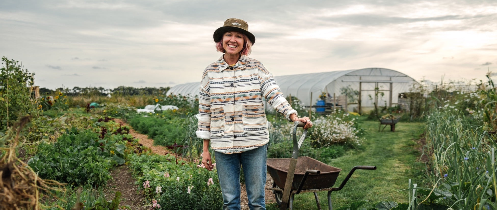 A woman stands with a spade by a wheelbarrow surrounded by rows of vegetables and plants