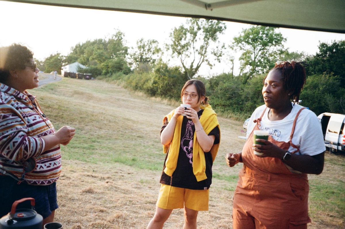 Three people standing outdoors under a canopy on a grassy field, with trees and a vehicle in the background. One person wears rust-colored overalls over a white shirt, another wears a black top with a yellow towel draped over the shoulders and yellow shorts, and a third person is partially visible on the left in a patterned fleece.
